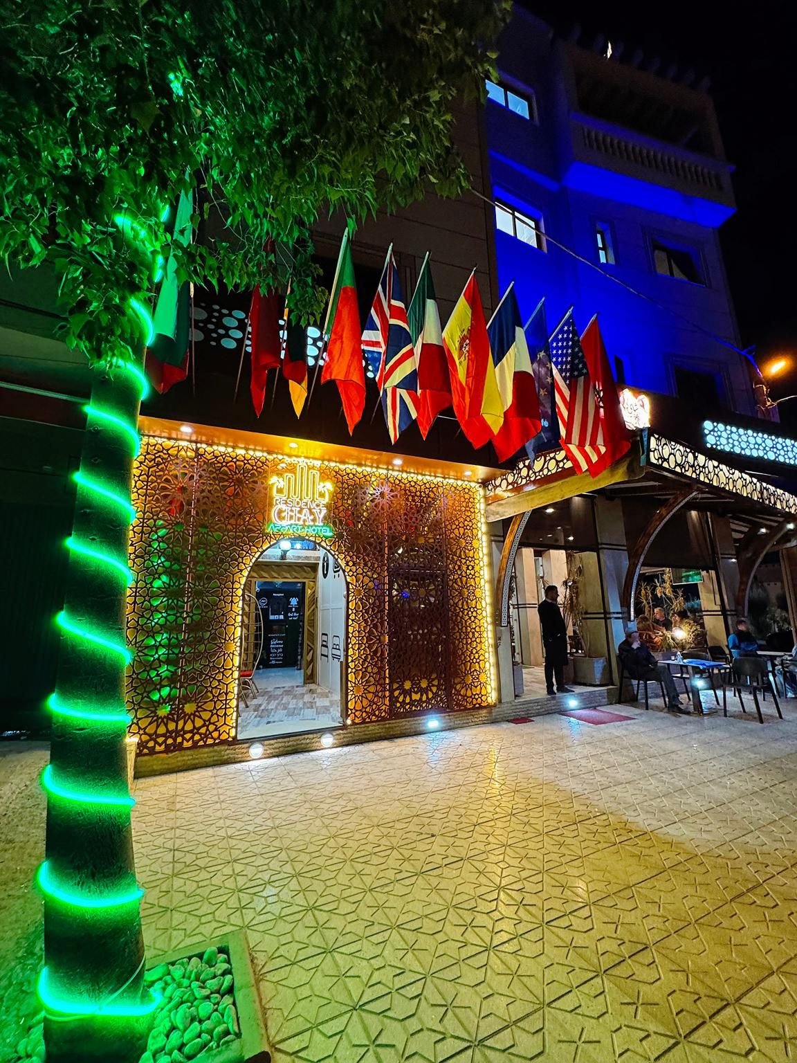 The entrance of Residence Chay at night: the lit lattice doorway, the neon sign and a row of flags on Hay El Massira